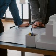 Close up of colleagues working on urban design with blueprints and building model. Team of people doing teamwork on construction layout, designing architectural development project.